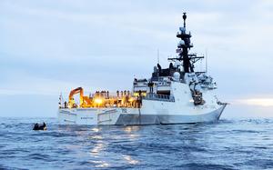 A U.S. Coast Guard cutter named Stratton (hull number 752) is illuminated by deck lights as it floats on calm ocean waters at dusk. Crew members are visible on the deck, and a small boat with personnel approaches the stern. The American flag flies atop the ship’s mast.