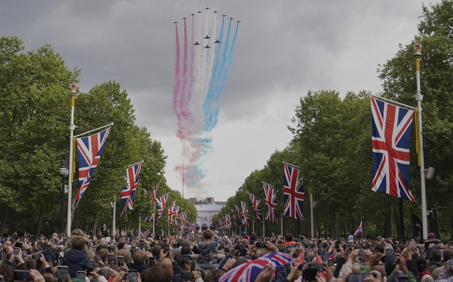 Military fighter jets, seen from below, emit red, white and blue smoke from their tails as they fly in formation over a large crowd of people and several poles adorned with the United Kingdom flag.