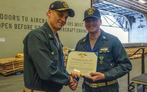 Two Navy officers in blue uniforms and hats shake hands and pose while holding a commendation award between them.