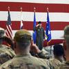 A military officer in flight suit salutes while standing at attention in front of four flags including the American flag and Air Force flags. The officer is positioned against a large American flag backdrop with red and white stripes. In the foreground, seated military personnel in camouflage uniforms and caps are saluting back.